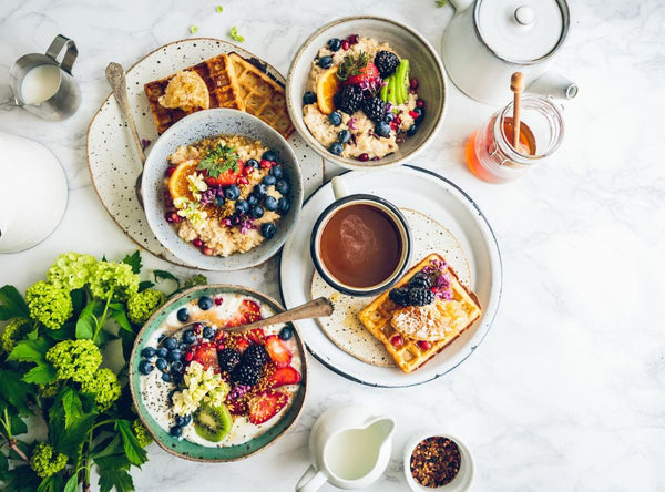 There are bowls and plates of breakfast food on a white marble table with green plants.